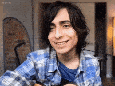 a young man in a blue plaid shirt is smiling while holding an acoustic guitar .