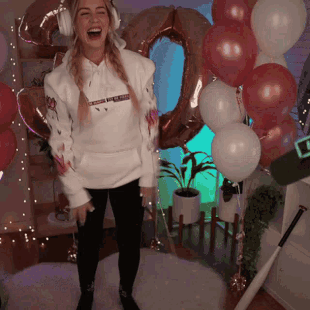 a woman wearing headphones and a hoodie with the number 20 on it laughs in front of balloons