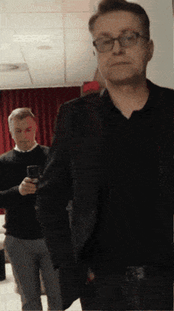 a man wearing glasses and a black jacket stands in front of a red curtain