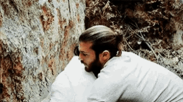 a man with long hair and a beard is kneeling down next to a rock .