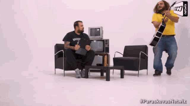 a man in a brooklyn shirt is sitting in a chair while another man plays a guitar