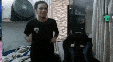a man in a black shirt is standing in a room with a fan and a chair .