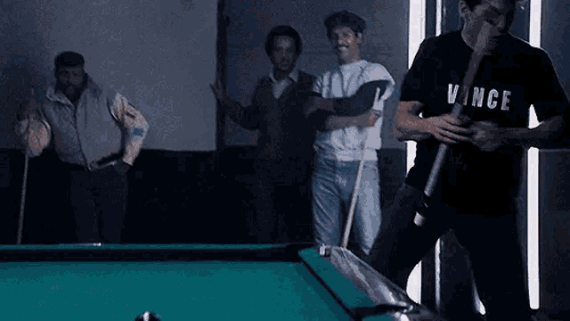 a group of men are playing pool in a dark room and one of them is holding a pool cue .