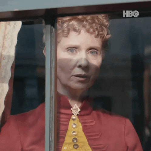 a woman in a red shirt and yellow tie looks out a window with hbo written on the bottom