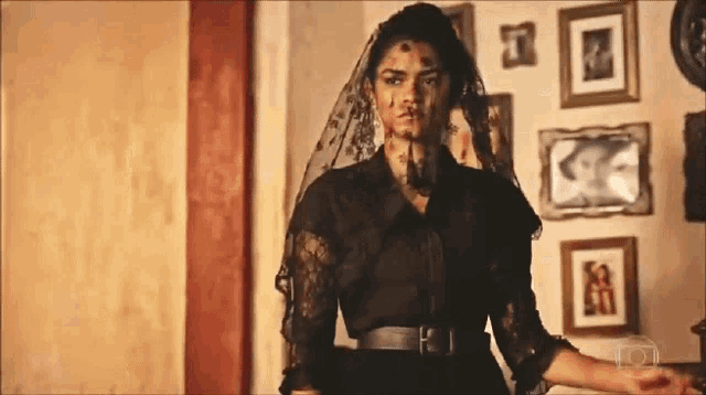a woman in a black dress with a veil on her face is standing in front of a wall with pictures on it .