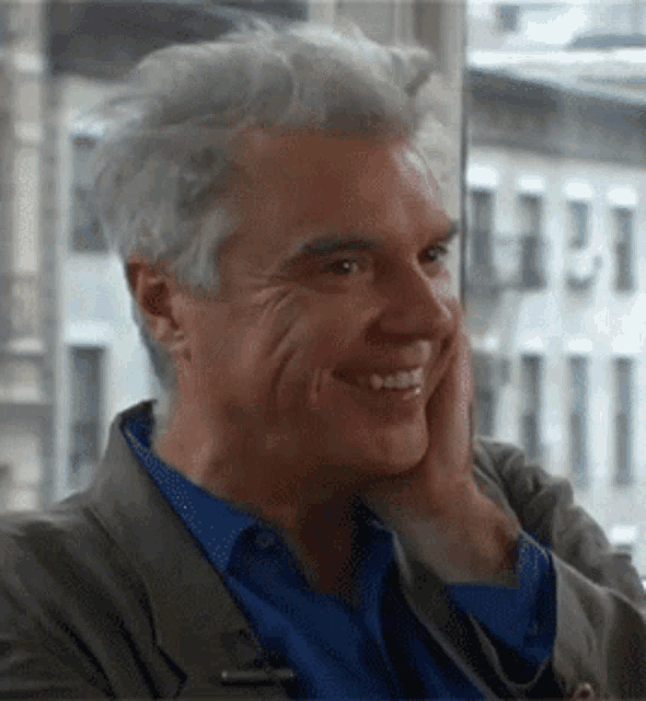 a man with gray hair and a blue shirt is smiling with his hand on his face