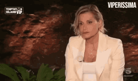 a woman in a white suit is sitting in front of a rock wall and talking to someone .