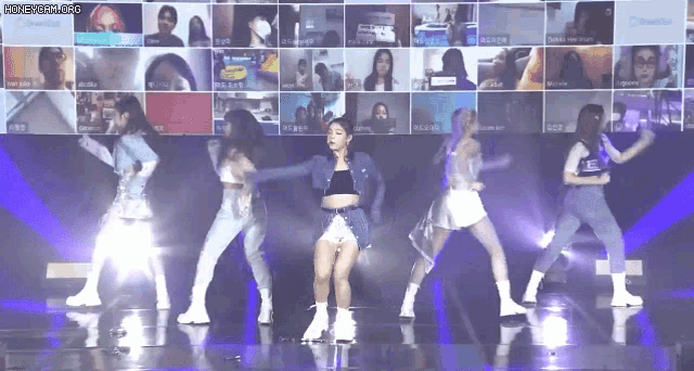 a group of women are dancing on a stage in front of a screen with a lot of people on it .