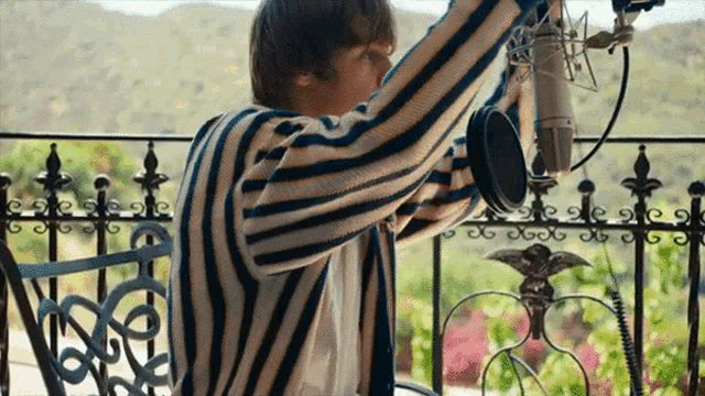 a man in a striped shirt is sitting on a balcony with a microphone in his hand .