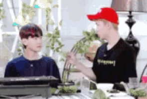a man in a red hat is talking to another man in a black shirt while sitting at a table .
