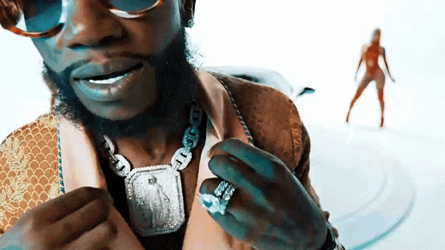 a close up of a man wearing a necklace and a ring with a lady in the background