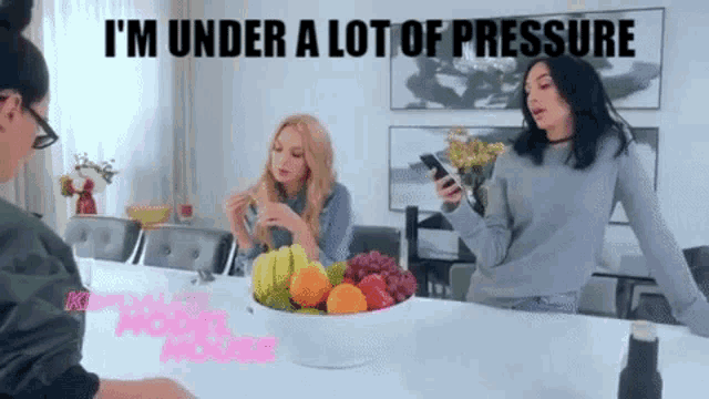 two women are sitting at a table with a bowl of fruit and a woman is holding a cell phone .