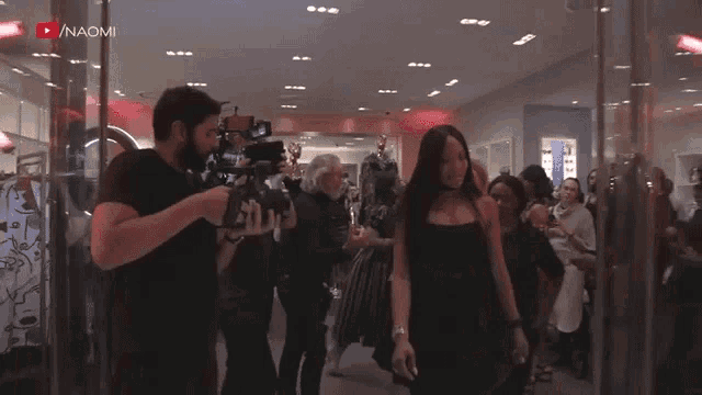 a woman in a black dress walks through a store with naom written on the bottom right