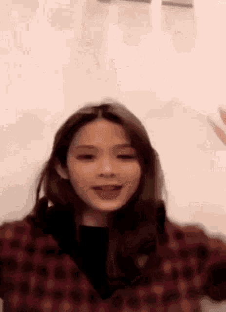 a woman in a plaid jacket is standing in front of a white wall and waving her hand .