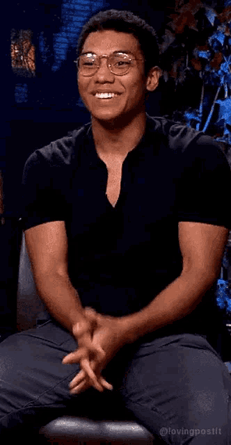 a man wearing glasses and a black shirt is smiling while sitting in a chair .