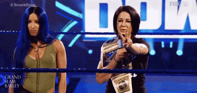 two women are standing in a wrestling ring one is holding a championship belt and pointing at the camera .