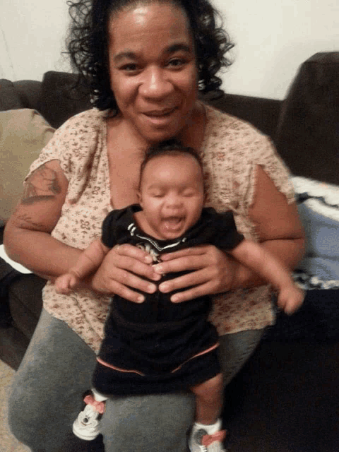 a woman sits on a couch holding a baby who is smiling