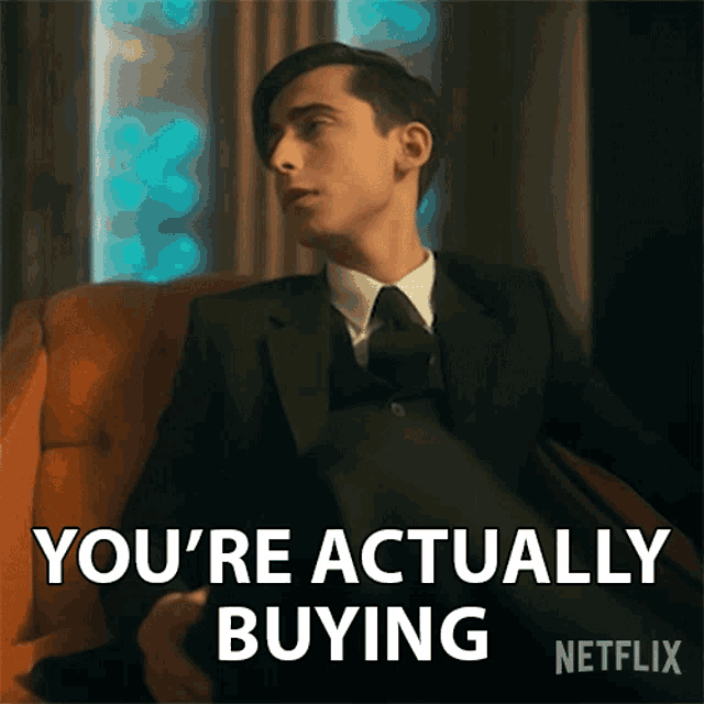 a man in a suit and tie is sitting in a chair and says you 're actually buying