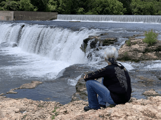 a woman in a black shirt with wings on the back sits on a rock near a waterfall