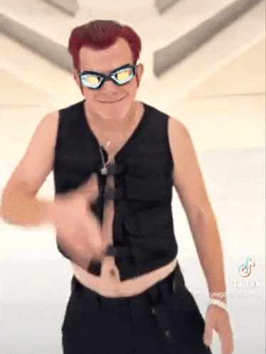 a man with red hair and sunglasses is wearing a black vest and black pants .