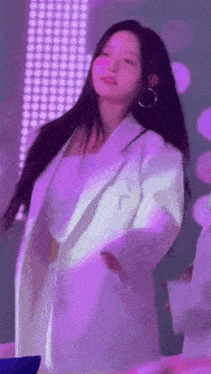 a woman in a white coat is dancing on a stage with purple lights behind her .