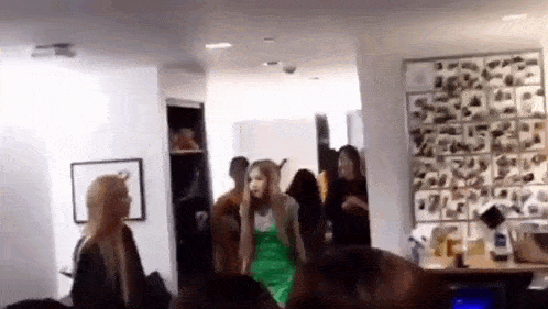 a group of people are standing in a room talking to each other . one of the women is wearing a green dress .