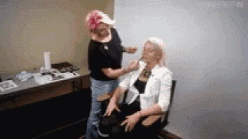 a woman is sitting in a chair getting her makeup done by a woman wearing a pink hat .
