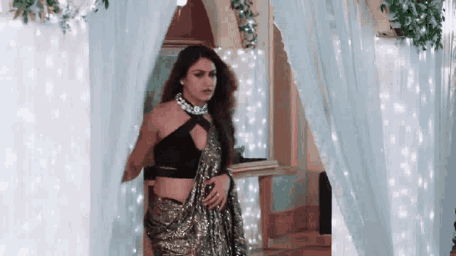 a woman in a black top and a gold saree is standing in front of a blue curtain .