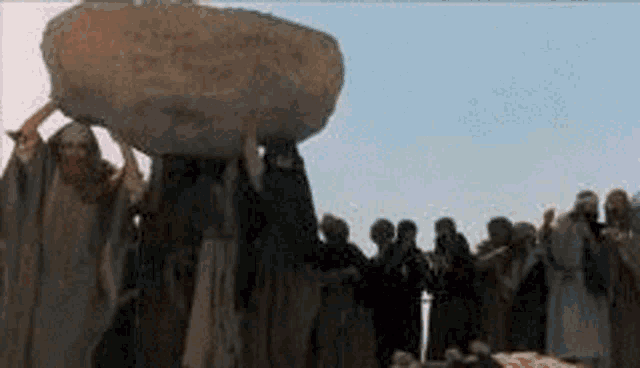 a man is carrying a large rock on his shoulders while a group of people watch .