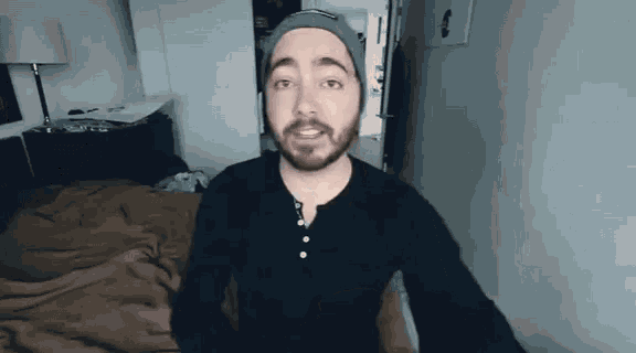 a man with a beard wearing a beanie is standing in front of a bed in a bedroom .