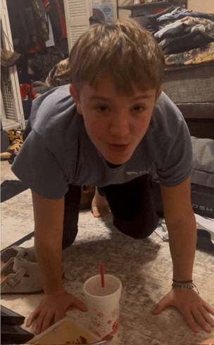 a boy in a blue shirt is crawling on the floor next to a cup with a red straw