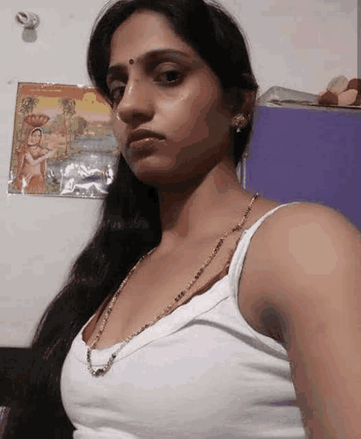 a woman in a white tank top is taking a selfie in front of a picture on the wall .