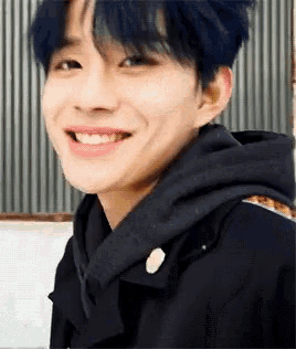 a young man with blue hair is smiling and wearing a black coat .