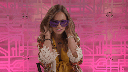 a woman wearing purple sunglasses stands in front of a pink wall