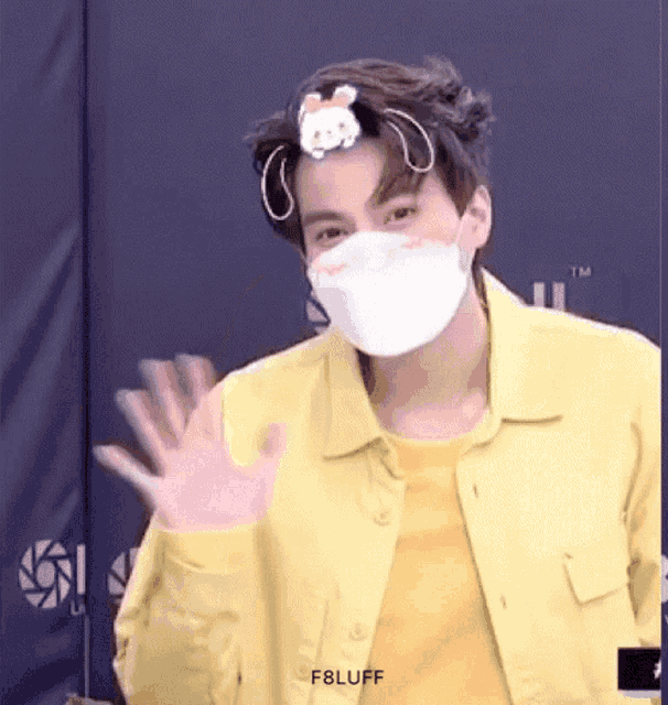 a young man wearing a yellow jacket and a white mask is waving his hand .