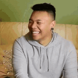 a man is sitting on a couch laughing and wearing a hoodie .
