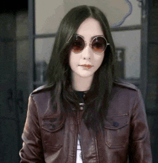 a woman wearing a leather jacket and round sunglasses is standing in front of a window .