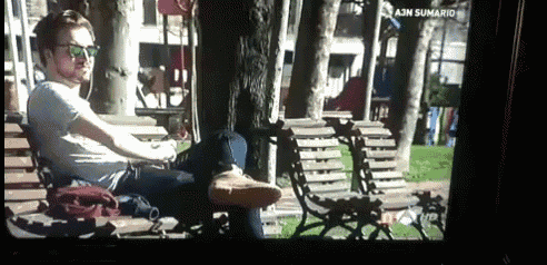 a man sits on a bench in a park with a screen that says ajn sumario on it