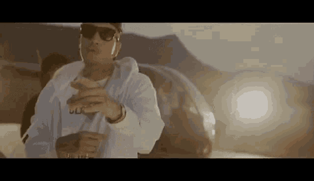 a man wearing sunglasses and a white hoodie is standing in front of a car in the desert .