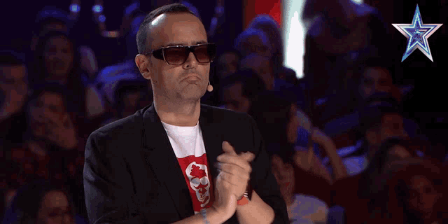 a man wearing sunglasses and a skull t-shirt applauds in front of a crowd