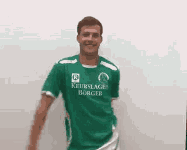 a man wearing a green shirt with the word keurslager on it is standing in front of a white wall .