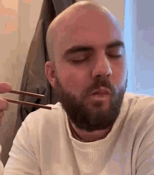 a bald man with a beard is eating with chopsticks and his eyes are closed .