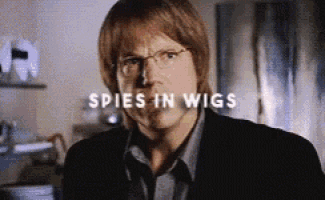 a close up of a man wearing glasses and a suit with the words spies in wigs behind him .