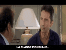 two men are talking to each other in front of a lamp and the words la classe mondiale are on the bottom of the screen .