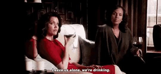 two women are sitting on a couch drinking wine and watching tv .