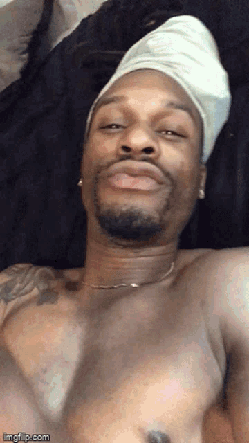 a shirtless man wearing a white towel on his head looks at the camera