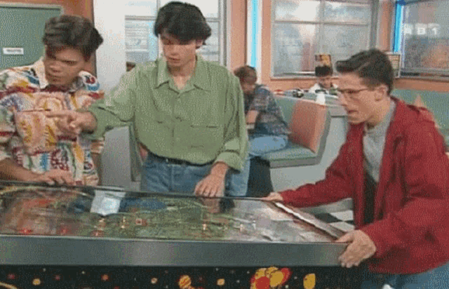 three men are playing a pinball machine in a diner with a sign that says ' tnt ' on it
