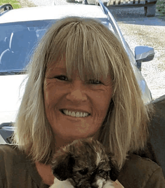 a woman is smiling while holding a small dog in front of a car