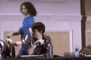 a man is sitting at a table with a microphone in front of him while another man stands behind him .