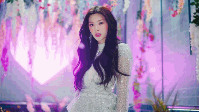 a woman in a white dress is standing in front of a purple background with flowers hanging from the ceiling .